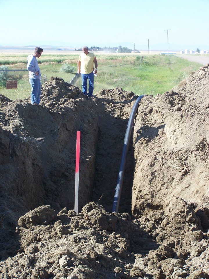 Two workers stand beside a deep trench with exposed pipe in a dirt field under a clear sky.