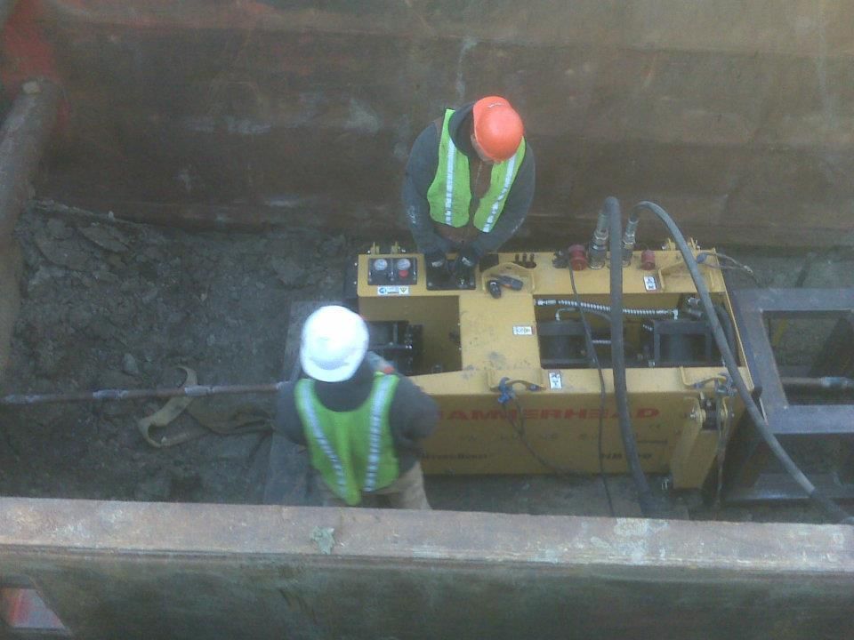 Two workers in hard hats inspect yellow construction equipment in a trench.