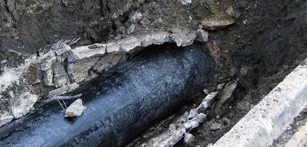 Black pipe exposed in a trench of dirt and concrete, with cracked surrounding ground