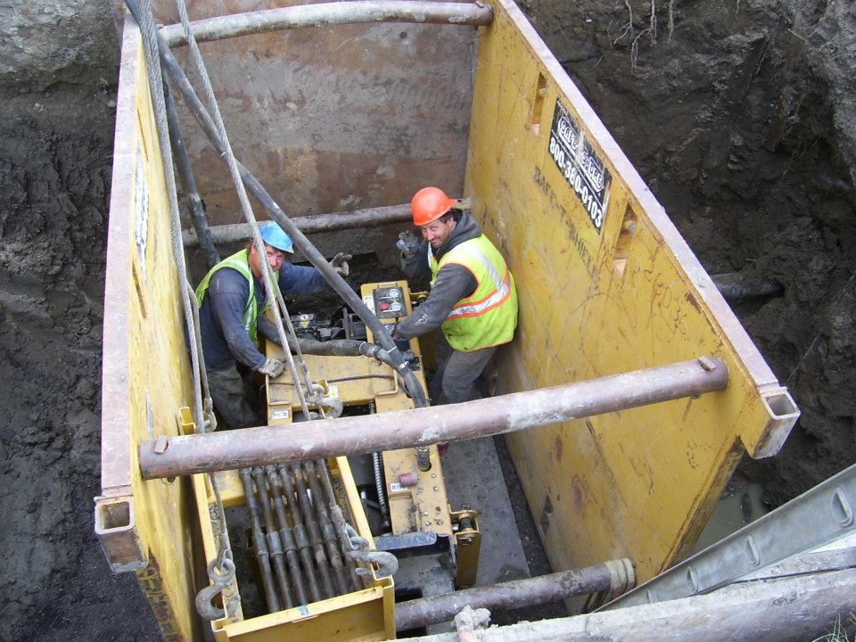 Workers in a deep trench operating construction equipment beside yellow shoring panels