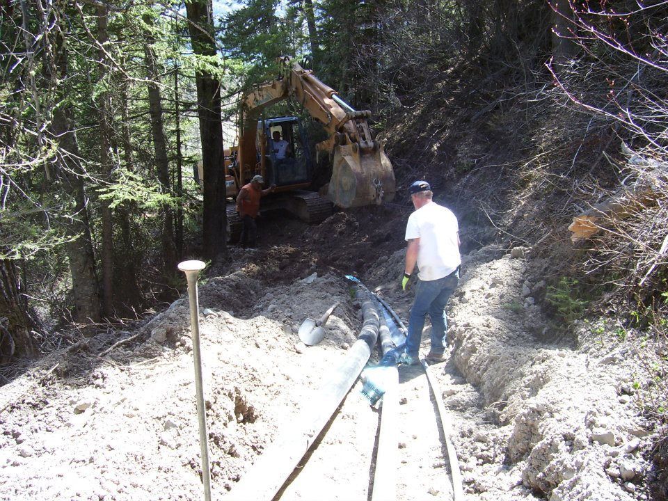 Man in white shirt stands in a muddy trench beside a backhoe in a wooded construction site.