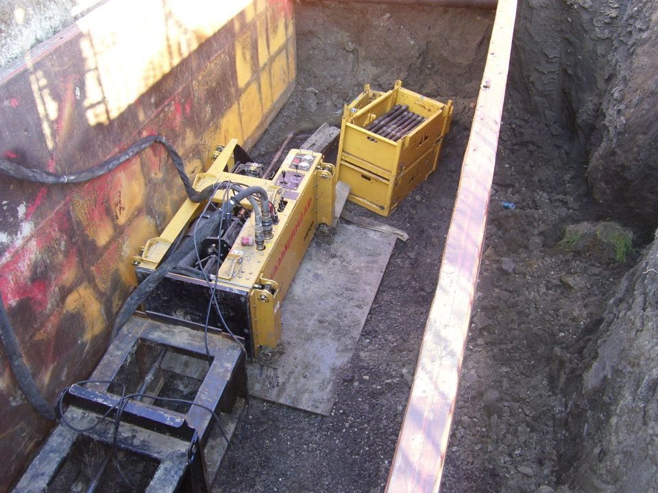 Construction equipment in a trench beside a graffiti-covered wall, with yellow machinery and a crate.