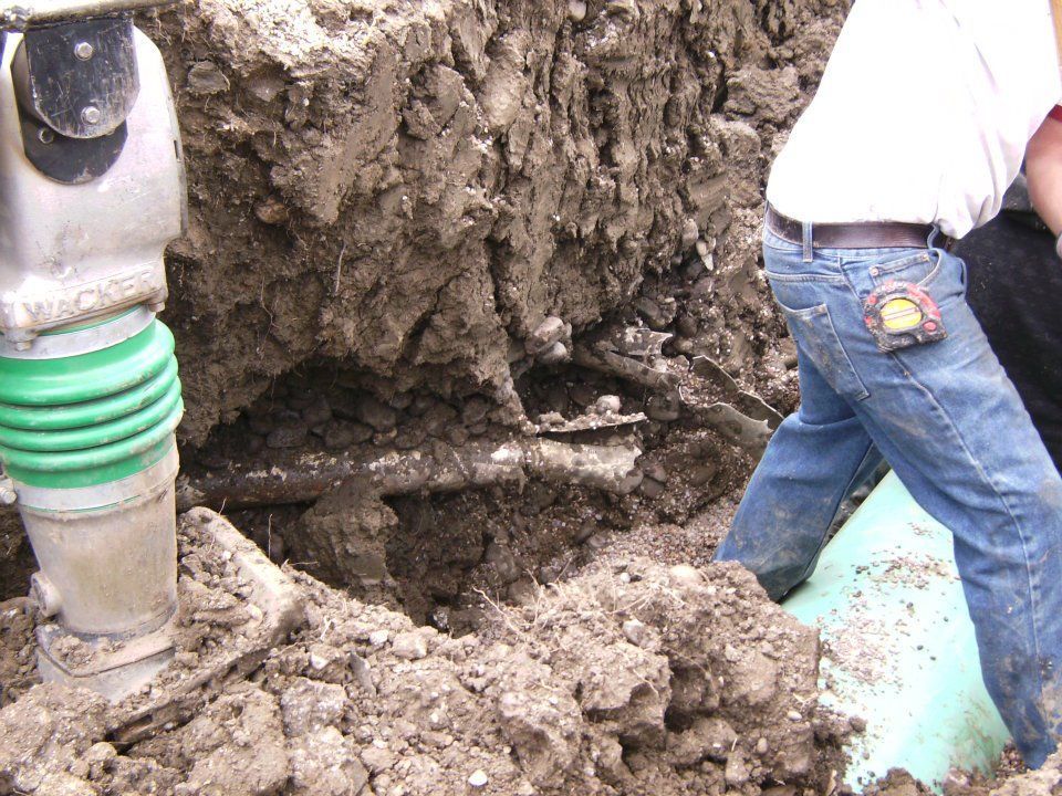 Workers digging a muddy trench around a broken pipe with heavy machinery nearby