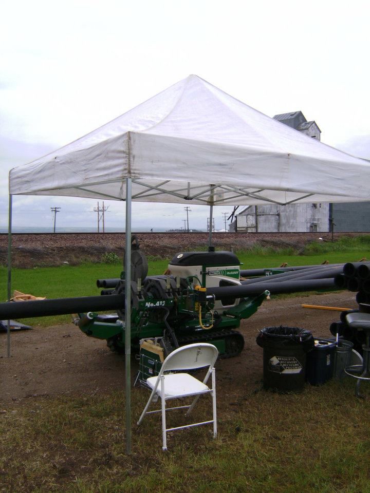 White canopy tent over farm equipment on a grassy field, with a folding chair in front