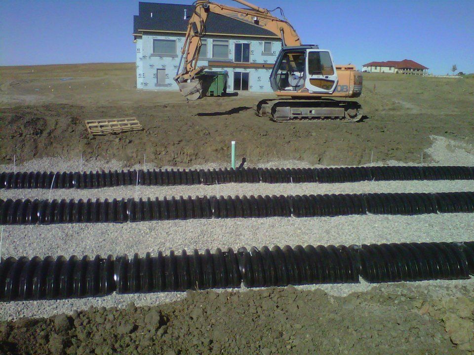 Excavator working beside a blue house on a muddy construction site with drainage pipes in the foreground
