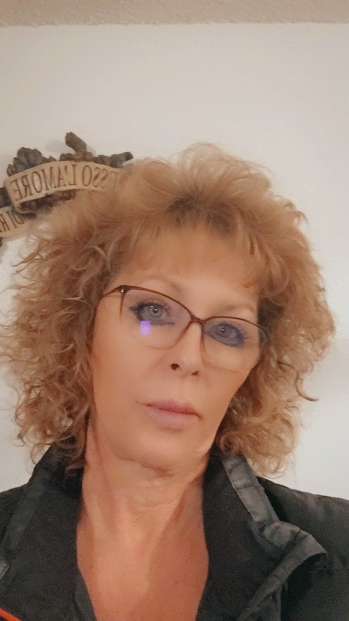 A woman wearing glasses and a black jacket is standing in front of a wall.
