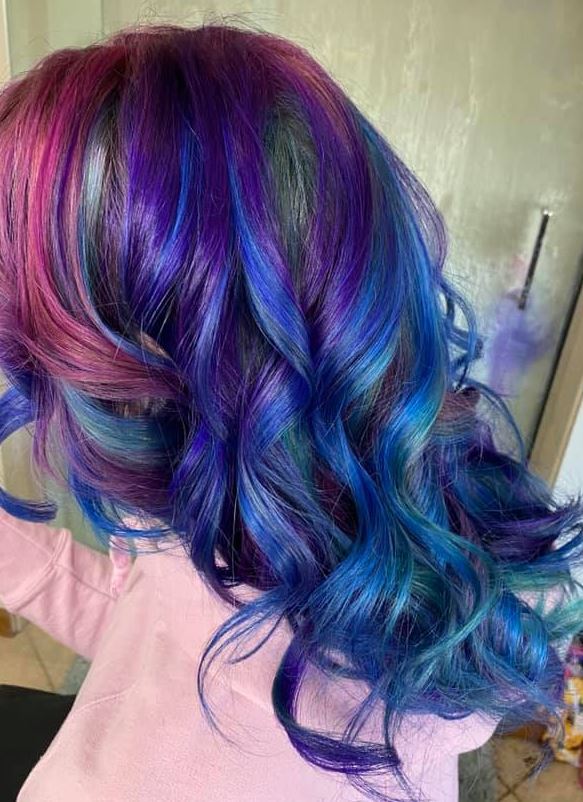 A woman with purple and blue hair is standing in front of a mirror.