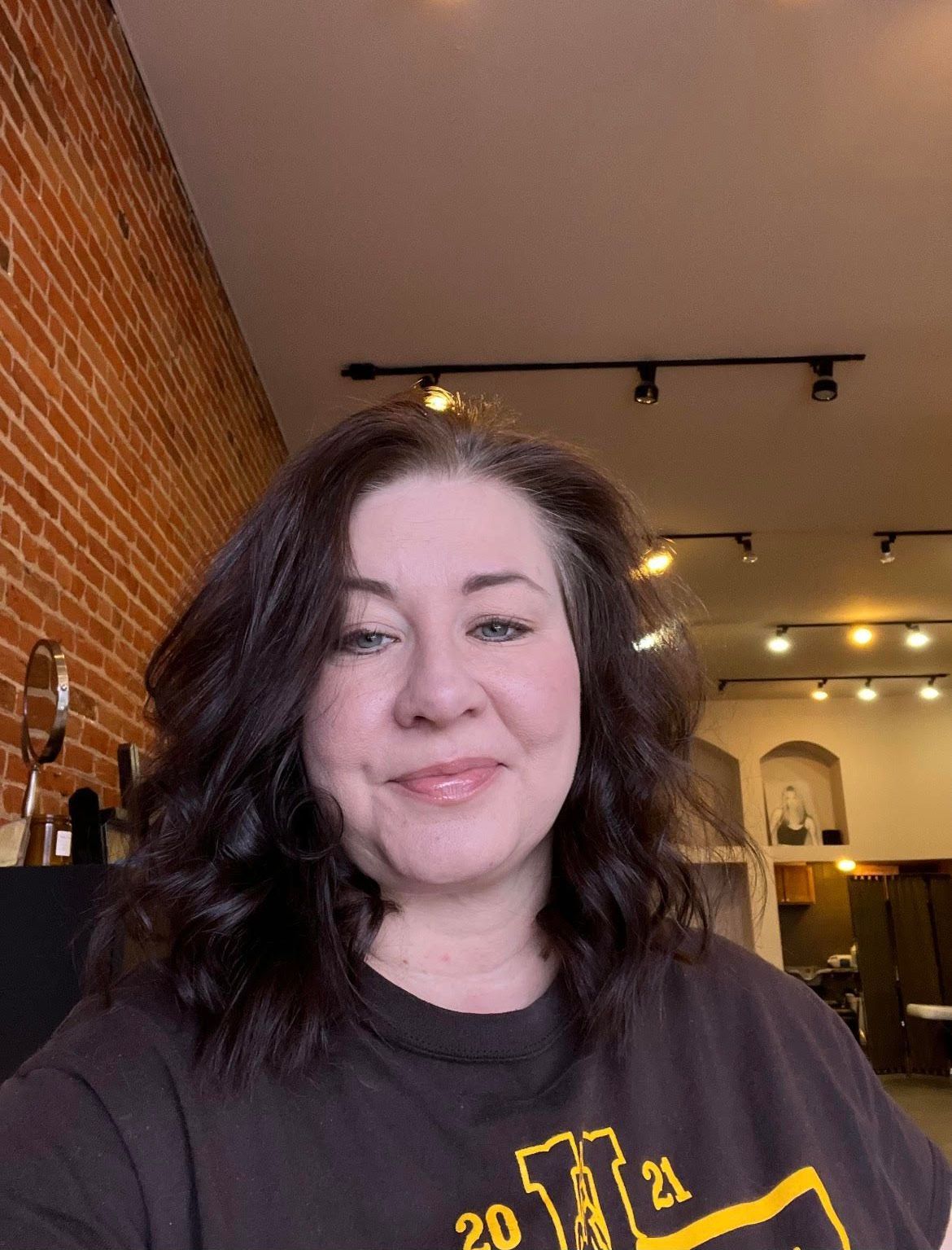 Woman with dark wavy hair smiles in a salon, brick wall background.