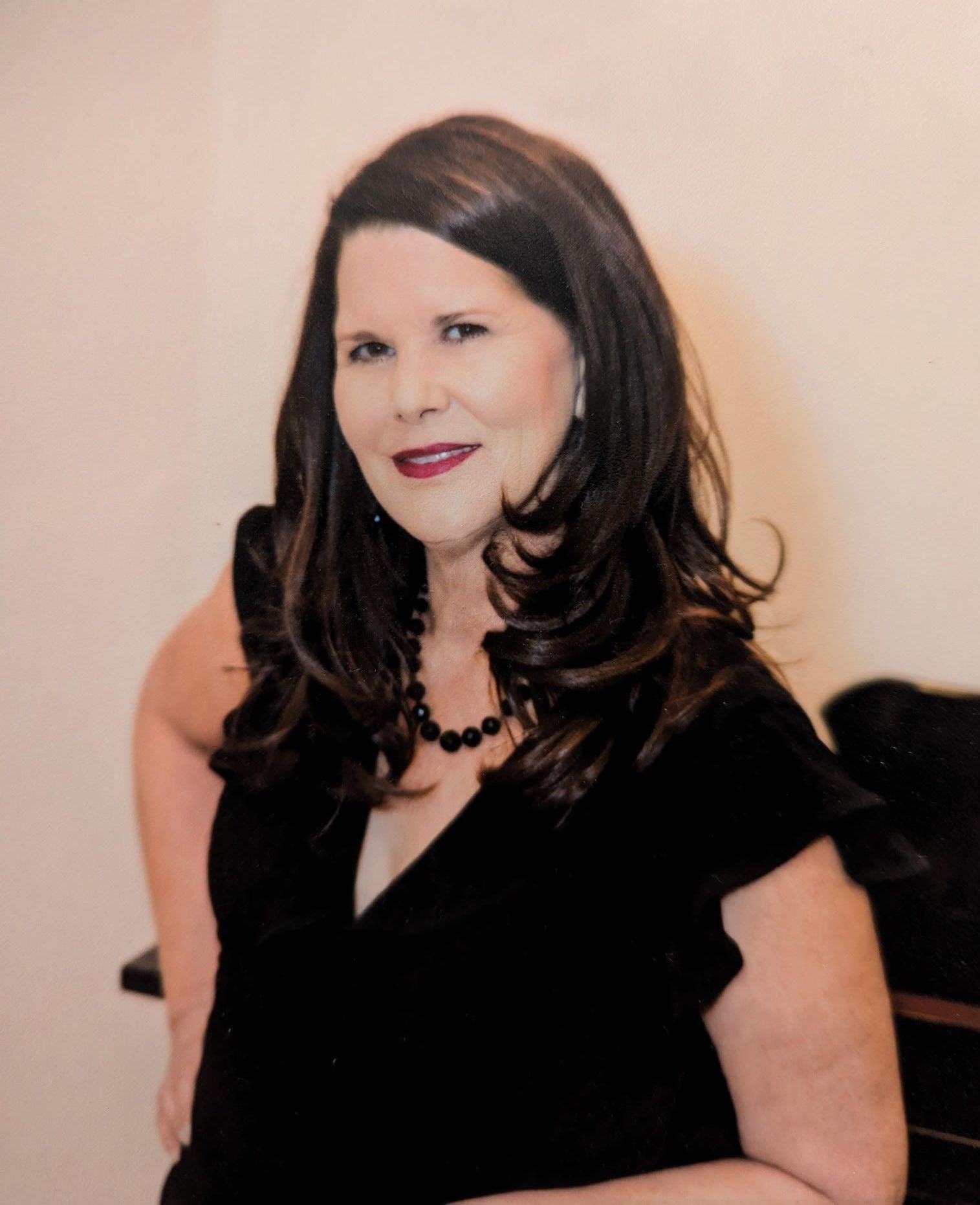 Woman with dark hair, wearing a black top and necklace, smiling. Arms resting, indoors.
