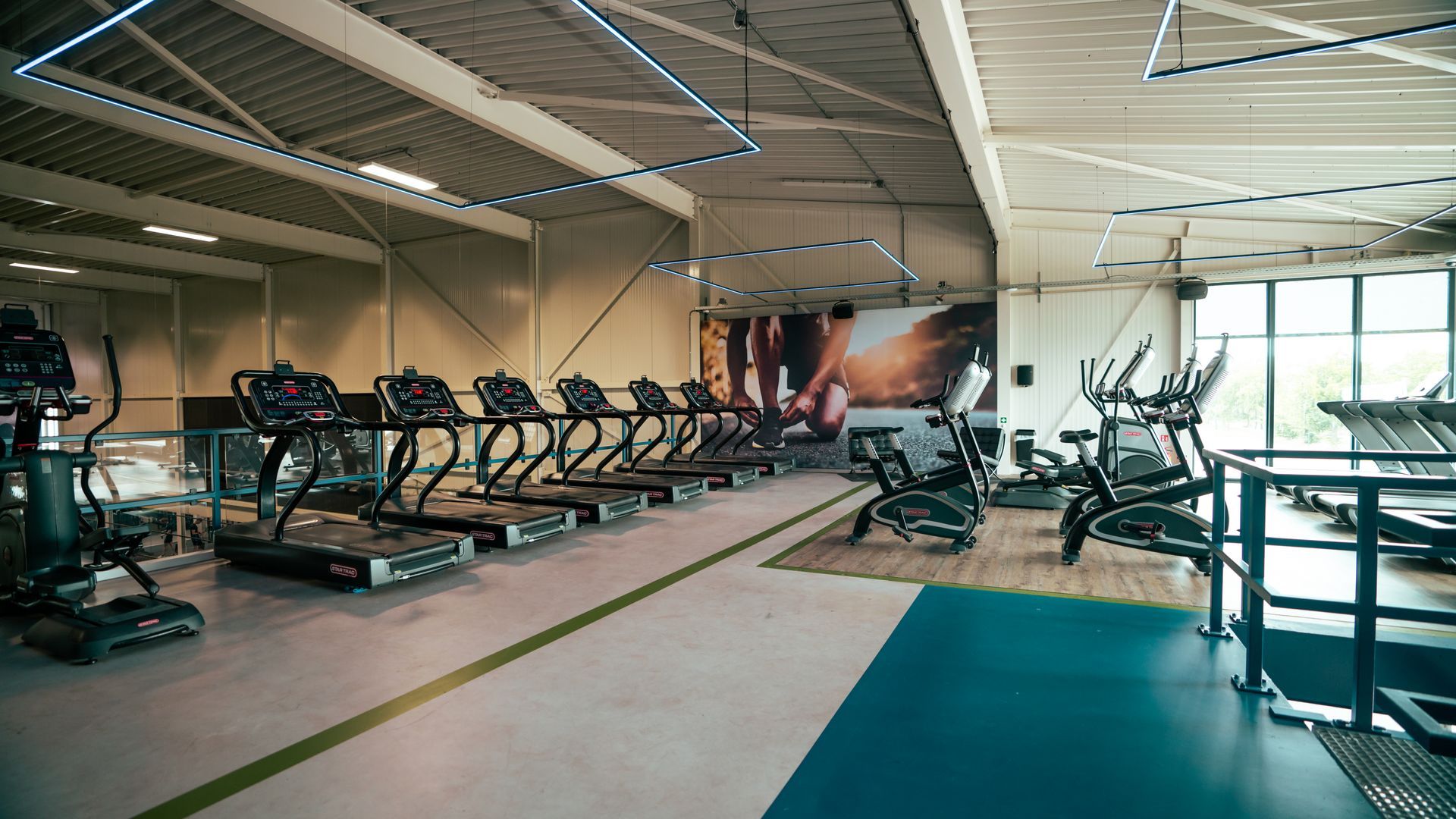 Binnengym met rijen loopbanden en hometrainers onder fel verlicht plafond.