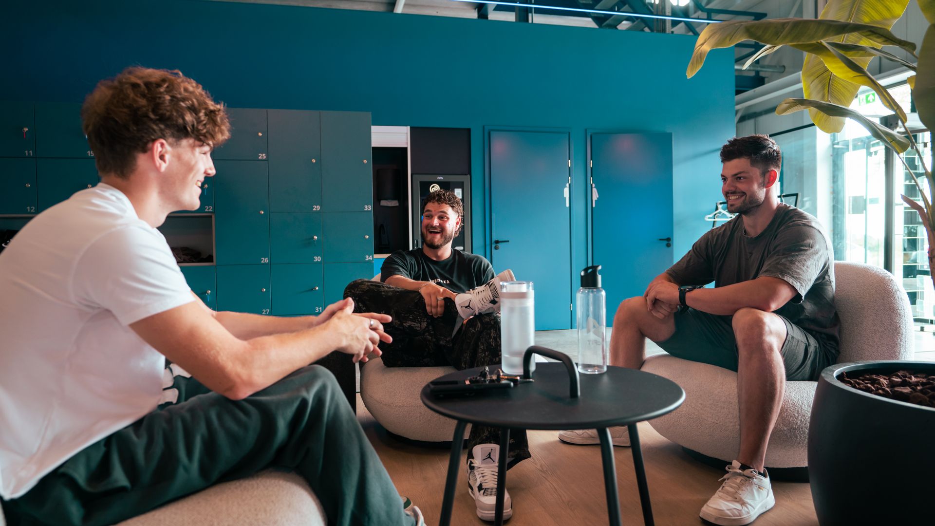 Drie mensen in gesprek in een moderne lounge met turquoise muren, een ronde tafel en potplanten.
