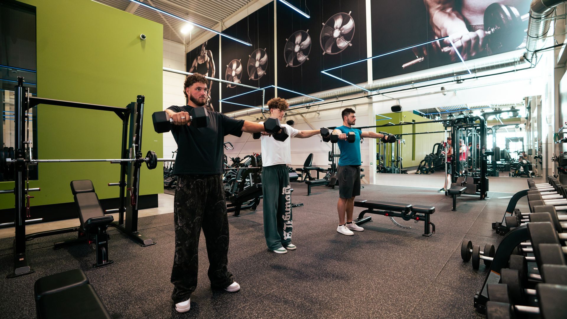 Drie mannen in een sportschool die dumbbeloefeningen doen, met de armen uitgestrekt.