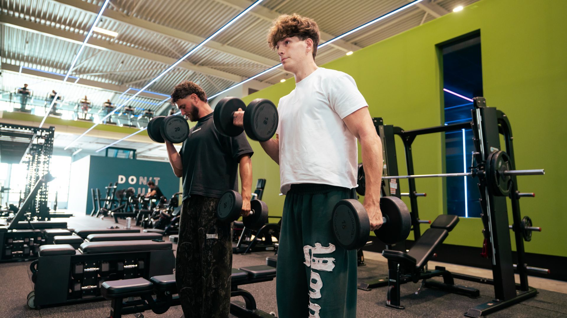 Twee mannen die dumbbells tillen in een sportschool. Groene en witte muren, fitnessapparatuur en gew