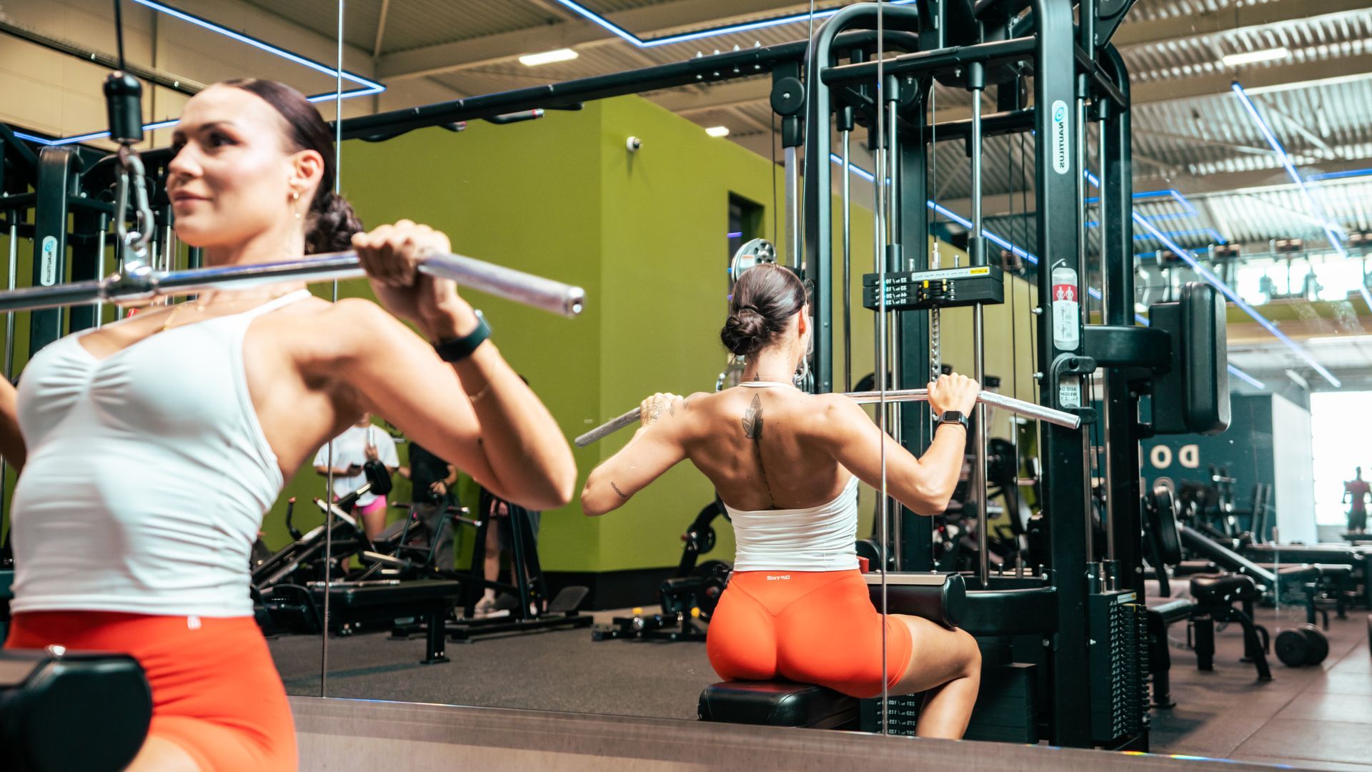 Twee mensen gebruiken gewichtsapparaten in een lichte sportschool, gekleed in witte shirts en oranje shorts.