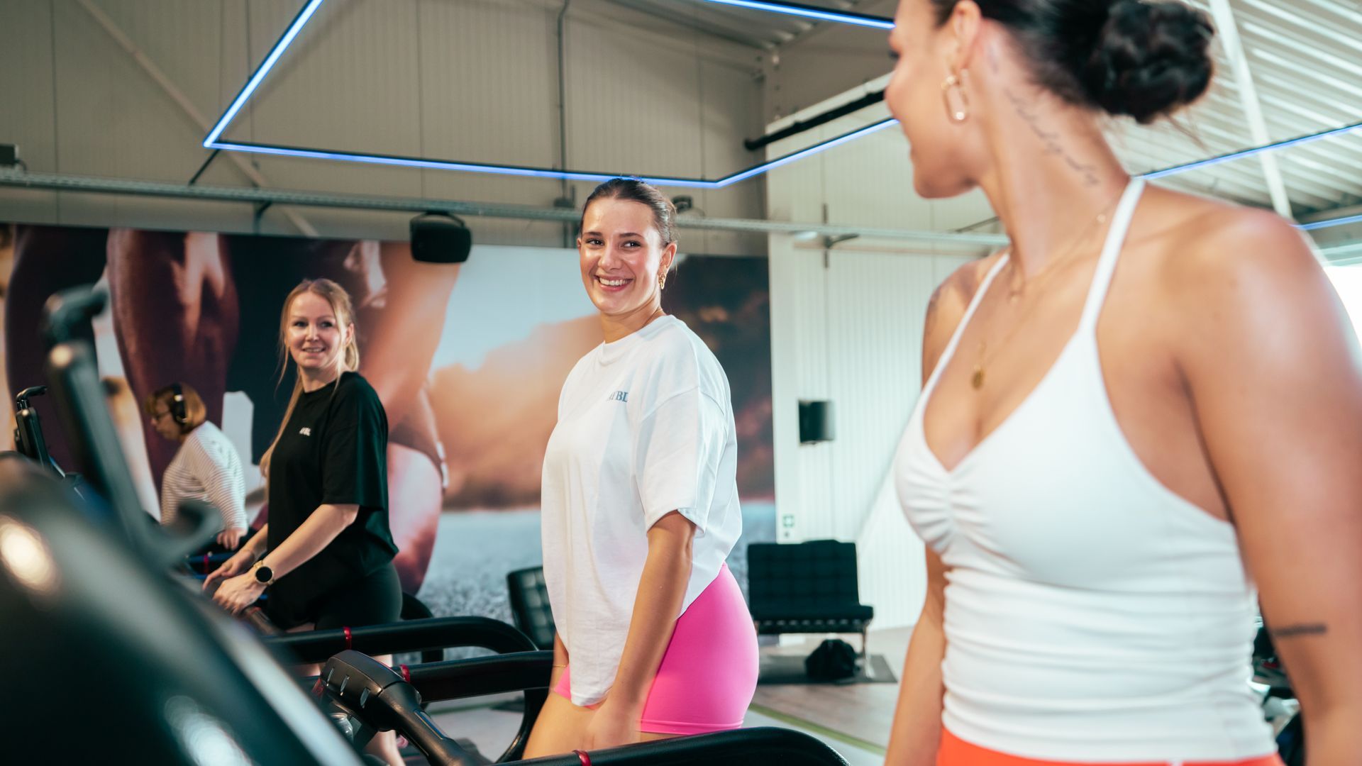 Drie vrouwen in sportkleding glimlachen in een lichte sportschool, vlakbij fitnessapparatuur.