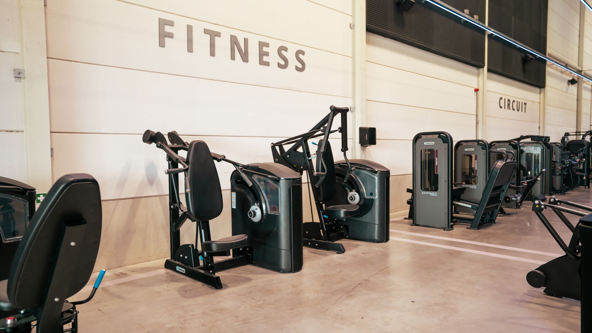 Sportschool met rijen zwarte fitnessapparaten onder een muur met het opschrift 