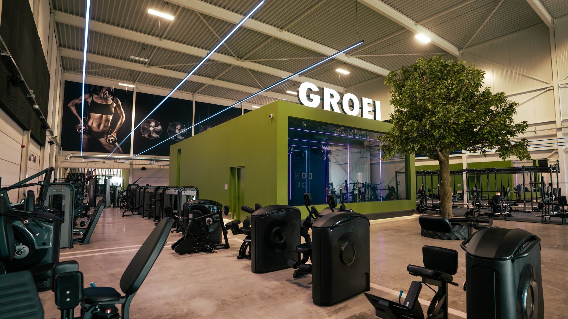 Twee mensen tillen halters in een sportschool, met spiegels, gewichten en industriële plafondbalken.