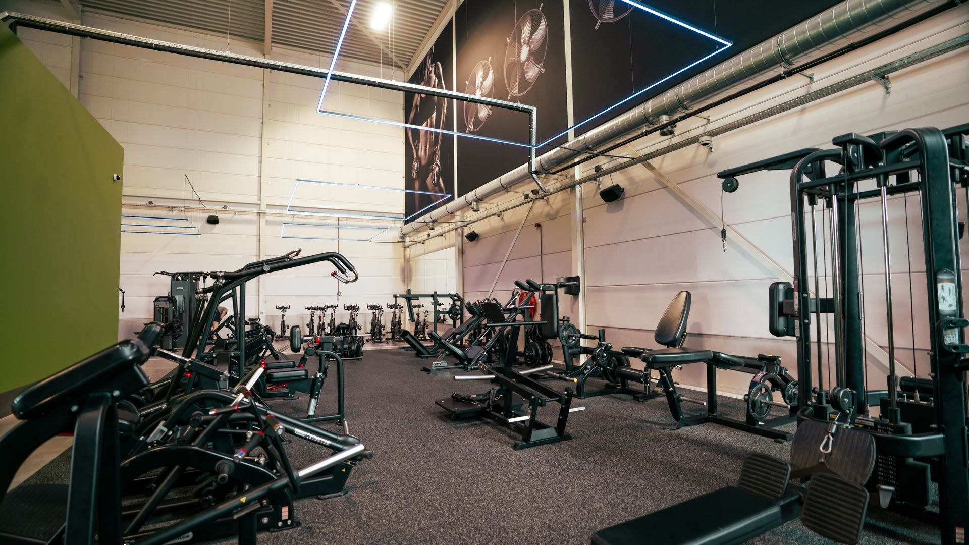 Een lege sportschool met gewichtsapparaten, banken en een zwarte vloer onder felle industriële lampen.