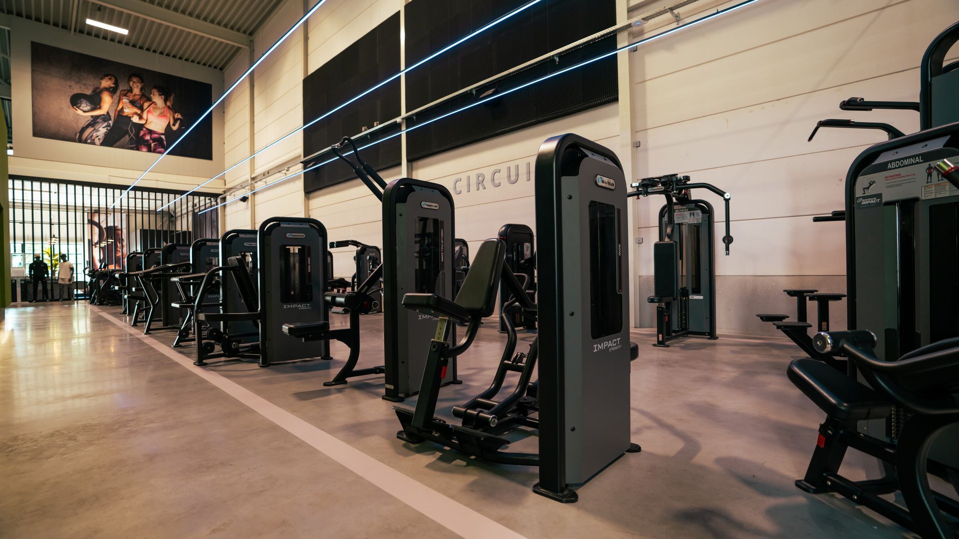 Modern sportschoolinterieur met rijen fitnessapparaten, beige muren en grote ramen met blauwe jaloezieën.