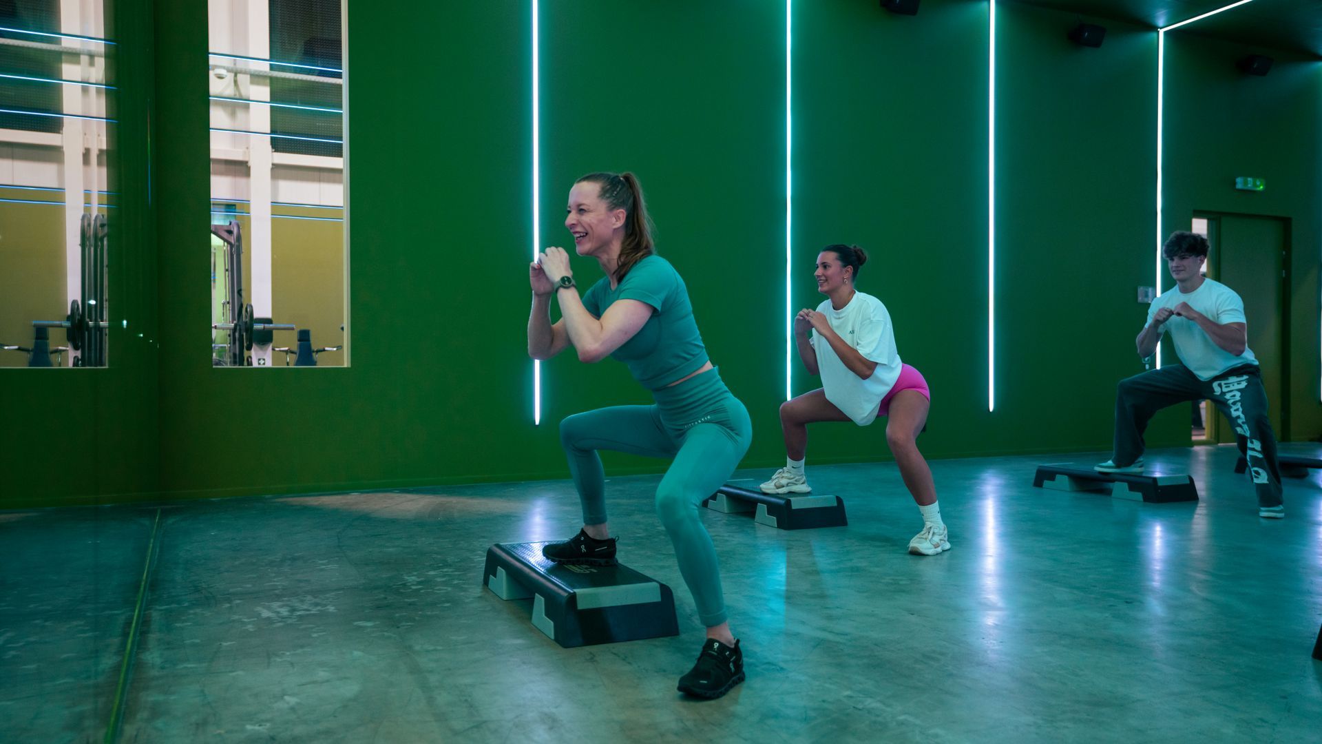 Mensen doen step-aerobics in een groen verlichte sportschool, waarbij ze gewichten vasthouden op verhoogde platforms.