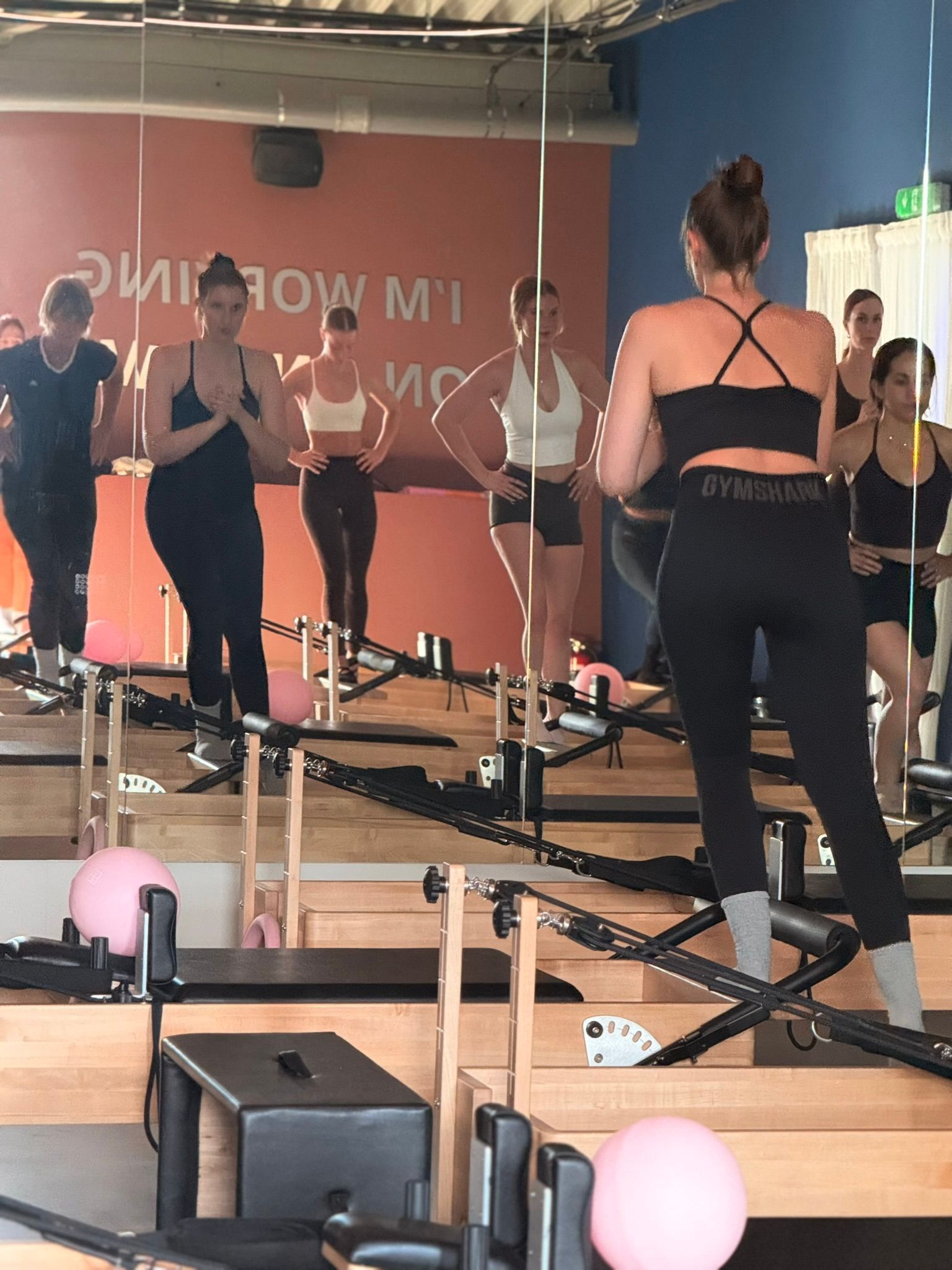 Mensen doen Pilates op reformers in een studio met spiegels en roze oefenballen.