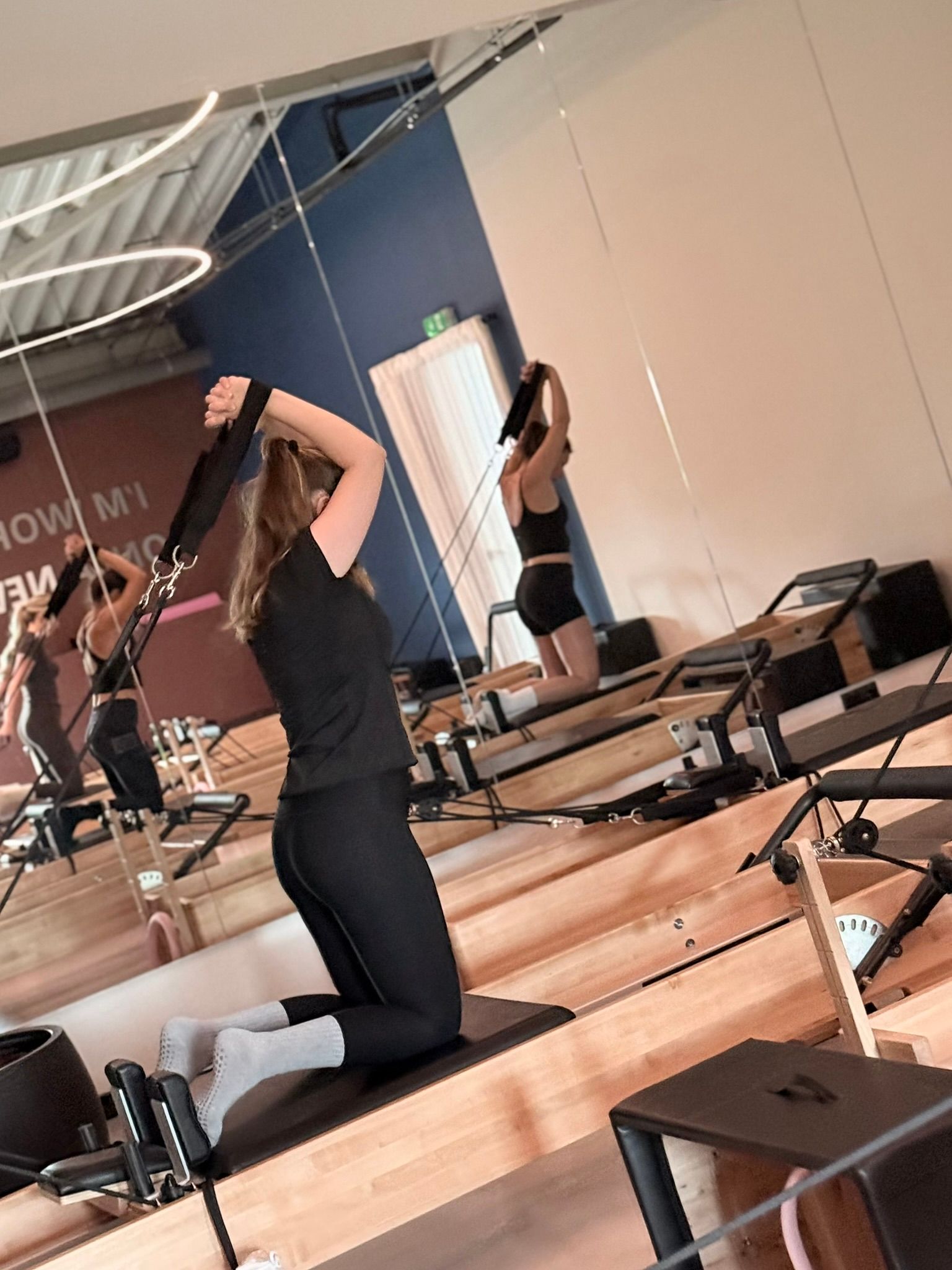 Een persoon knielt op een Pilates-reformer en reikt boven zijn hoofd in een studio met spiegelwanden.
