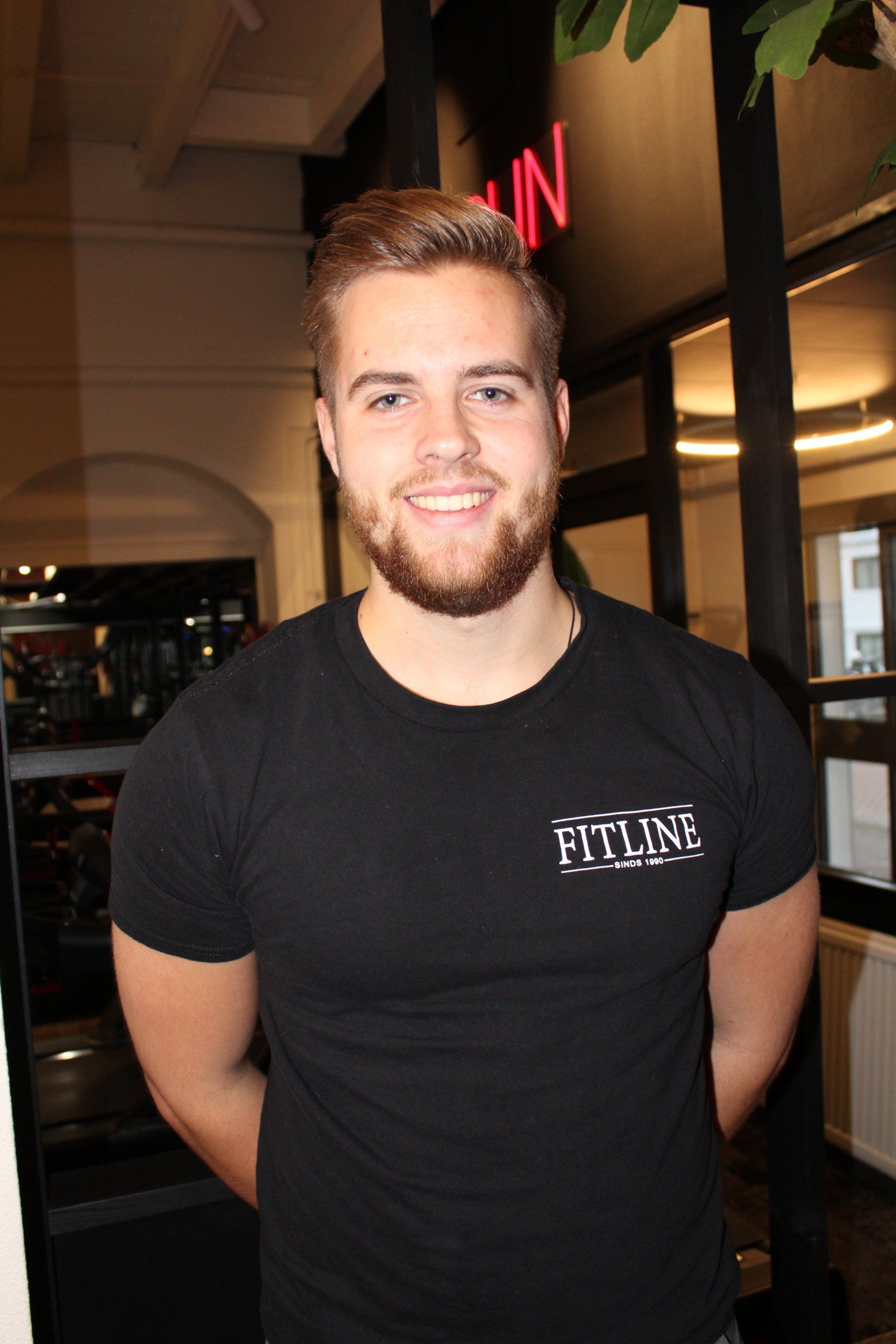 Een lachende man in een zwart T-shirt staat binnen bij een winkelraam.
