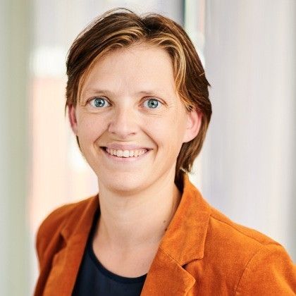 Een lachend persoon in een oranje blazer tegen een zacht vervaagde achtergrond.