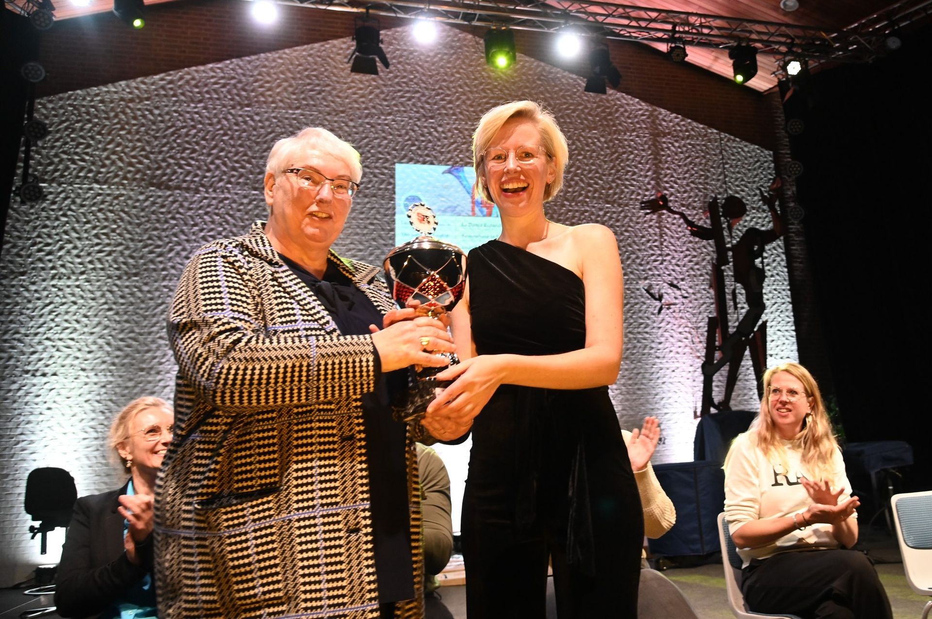 Twee vrouwen staan op het podium met een prijs in hun handen, met op de achtergrond zittende mensen en podiumverlichting.