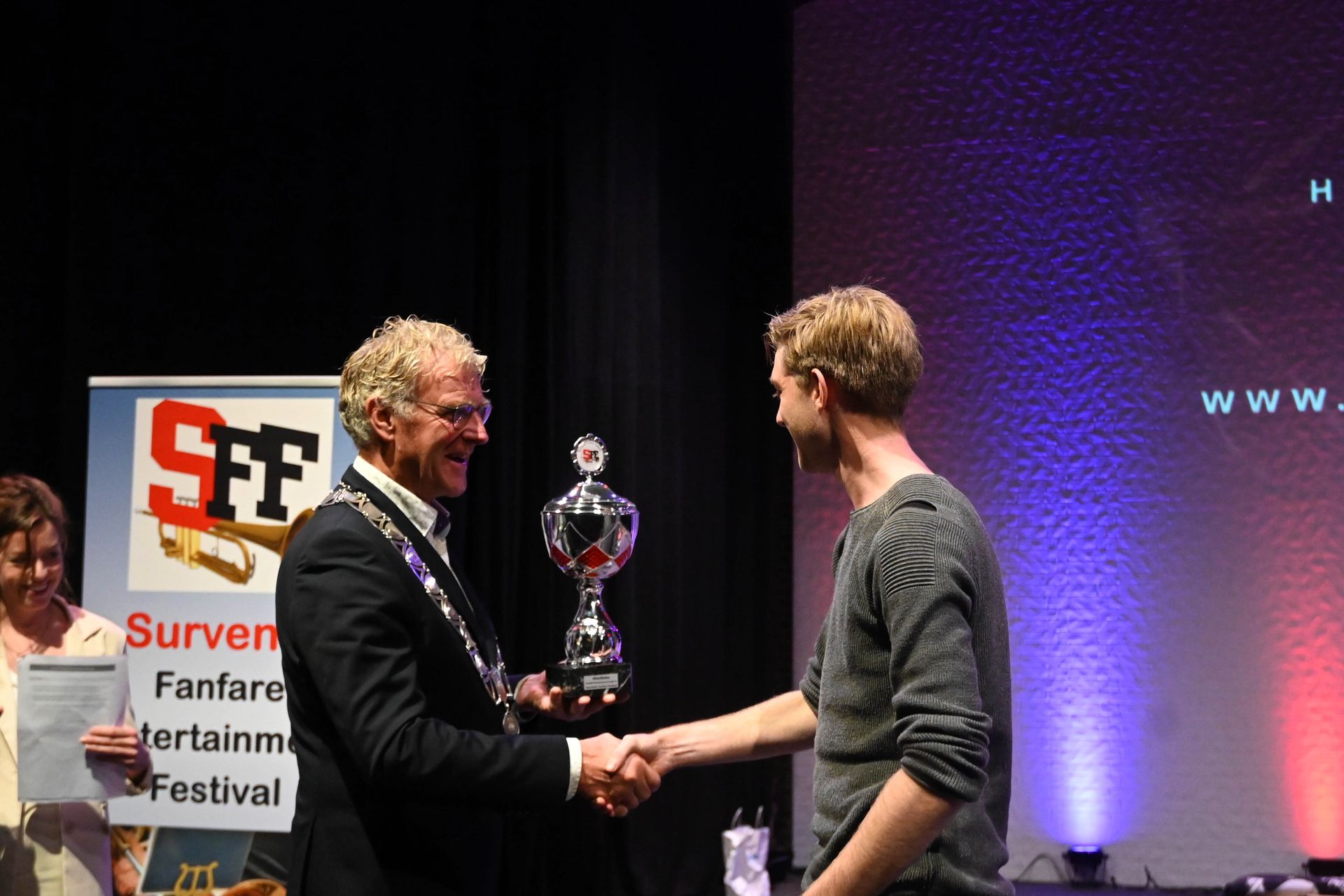 Een man ontvangt een trofee terwijl hij handen schudt op een podium met een spandoek van het Surviving Heritage Festival.