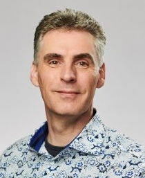 Portret van een lachende man in een overhemd met patroon tegen een grijze achtergrond.