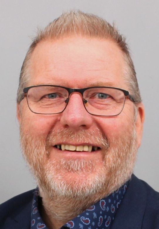 Een lachende man met een bril en een grijze baard, gekleed in een donkere blazer en een overhemd met een patroon.