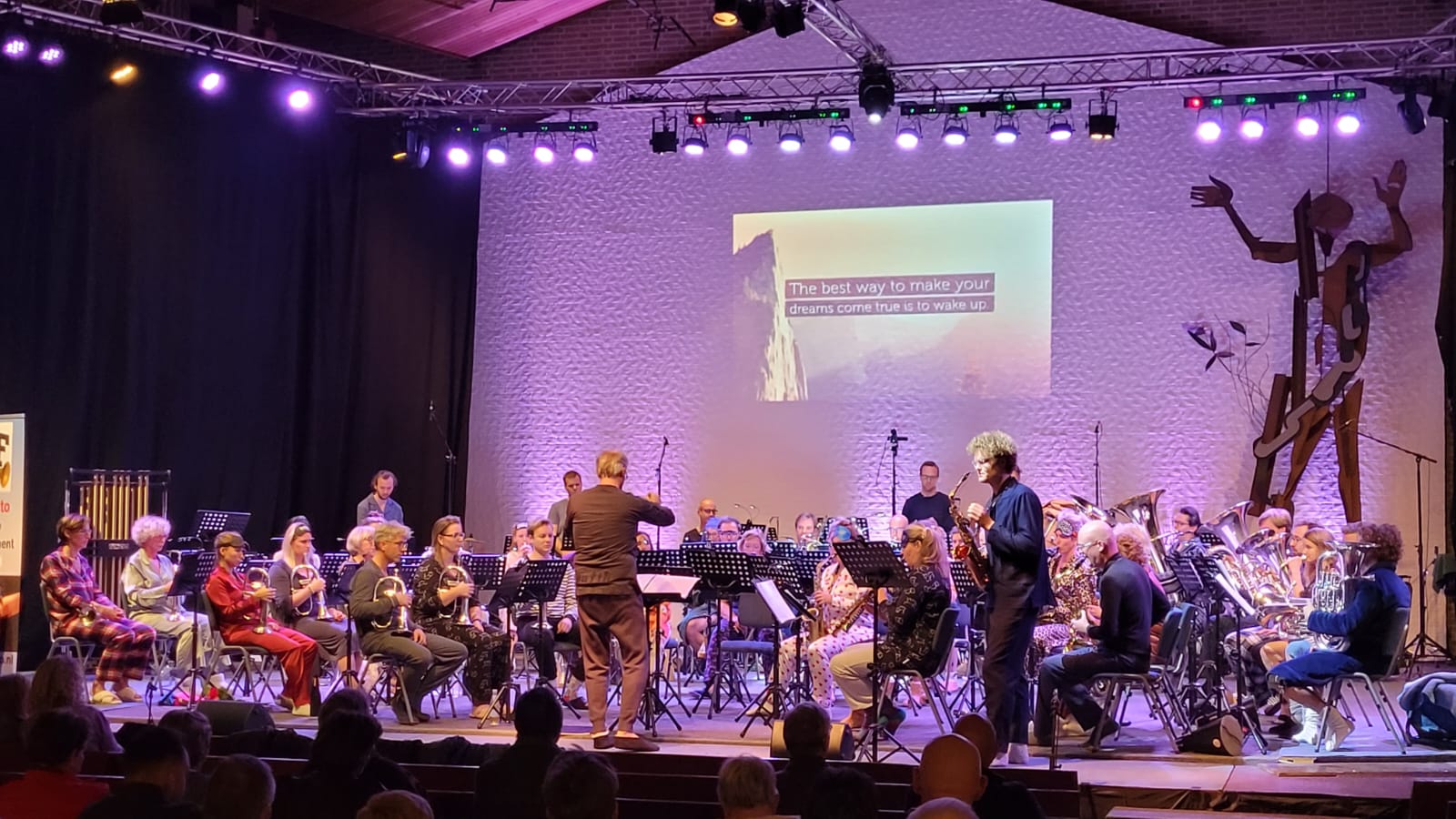 Orkest treedt op op een paars verlicht podium met een dirigent en een projectiescherm als achtergrond.