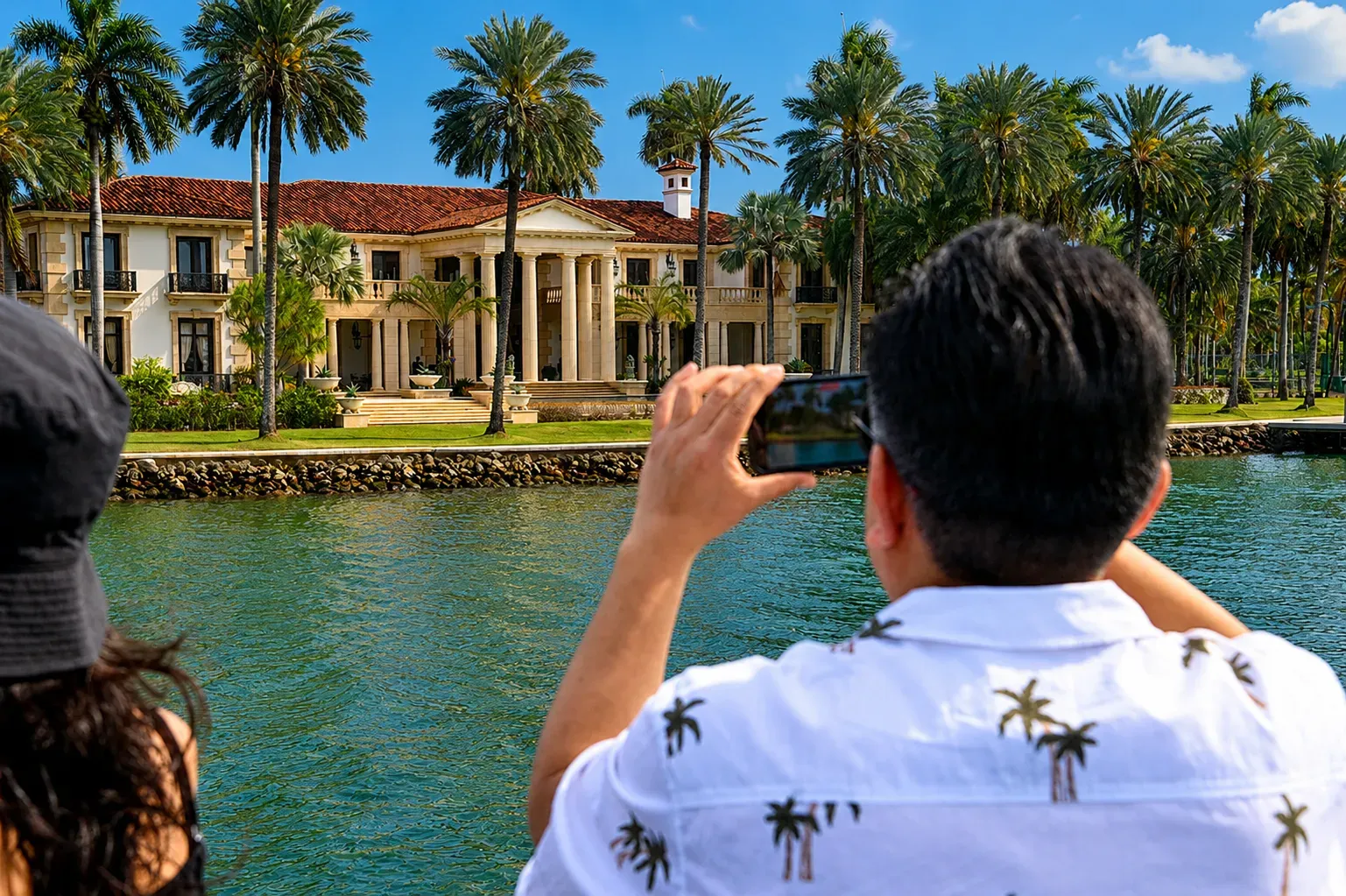 Visitor photographing a luxury waterfront mansion on Star Island Miami from Biscayne Bay surrounded by palm trees and celebrity homes