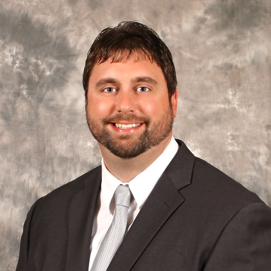 Headshot of a smiling man in a dark suit and silver tie against a gray studio backdrop
