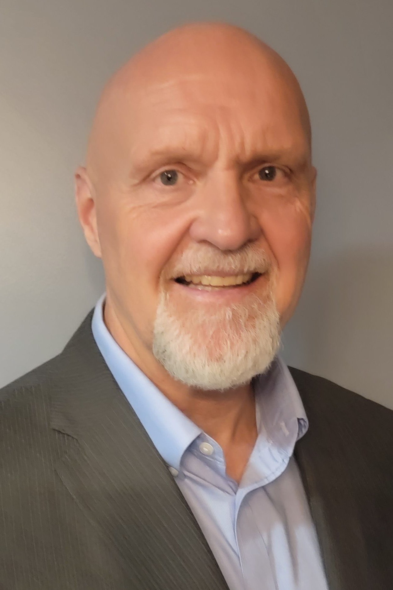 Smiling bald man with a white goatee in a gray suit and blue shirt against a neutral background