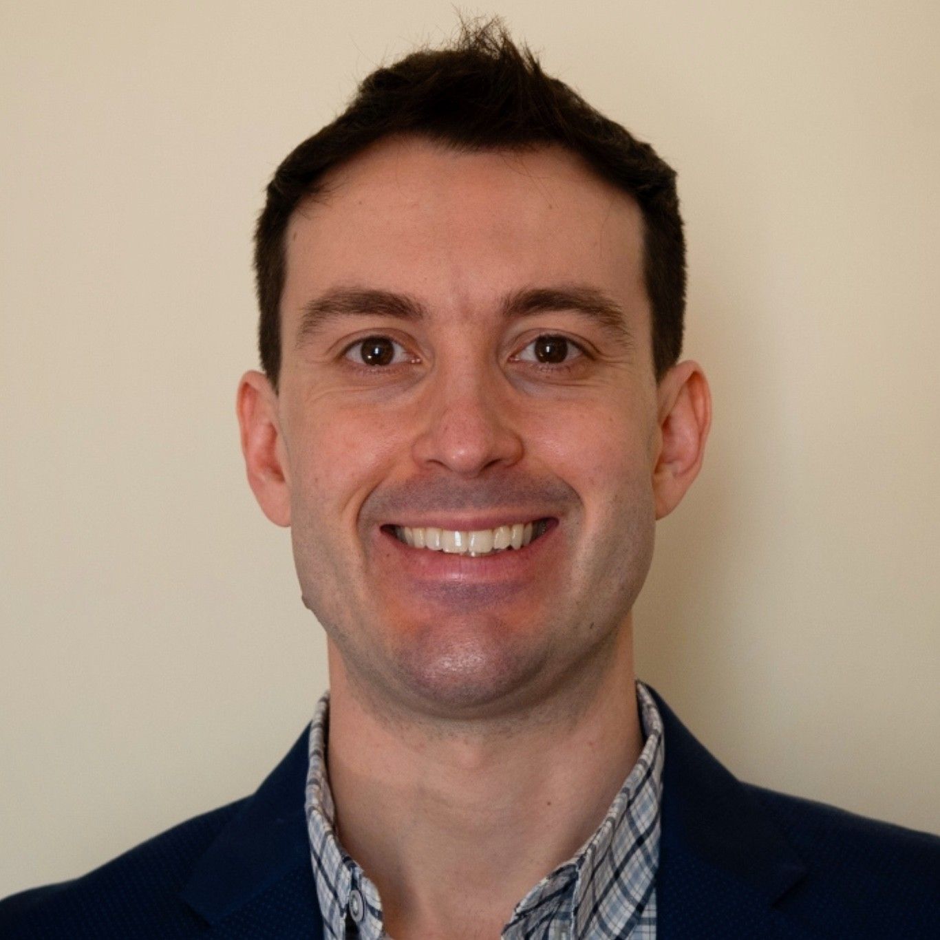 Smiling man in a navy blazer and checkered shirt against a plain beige background