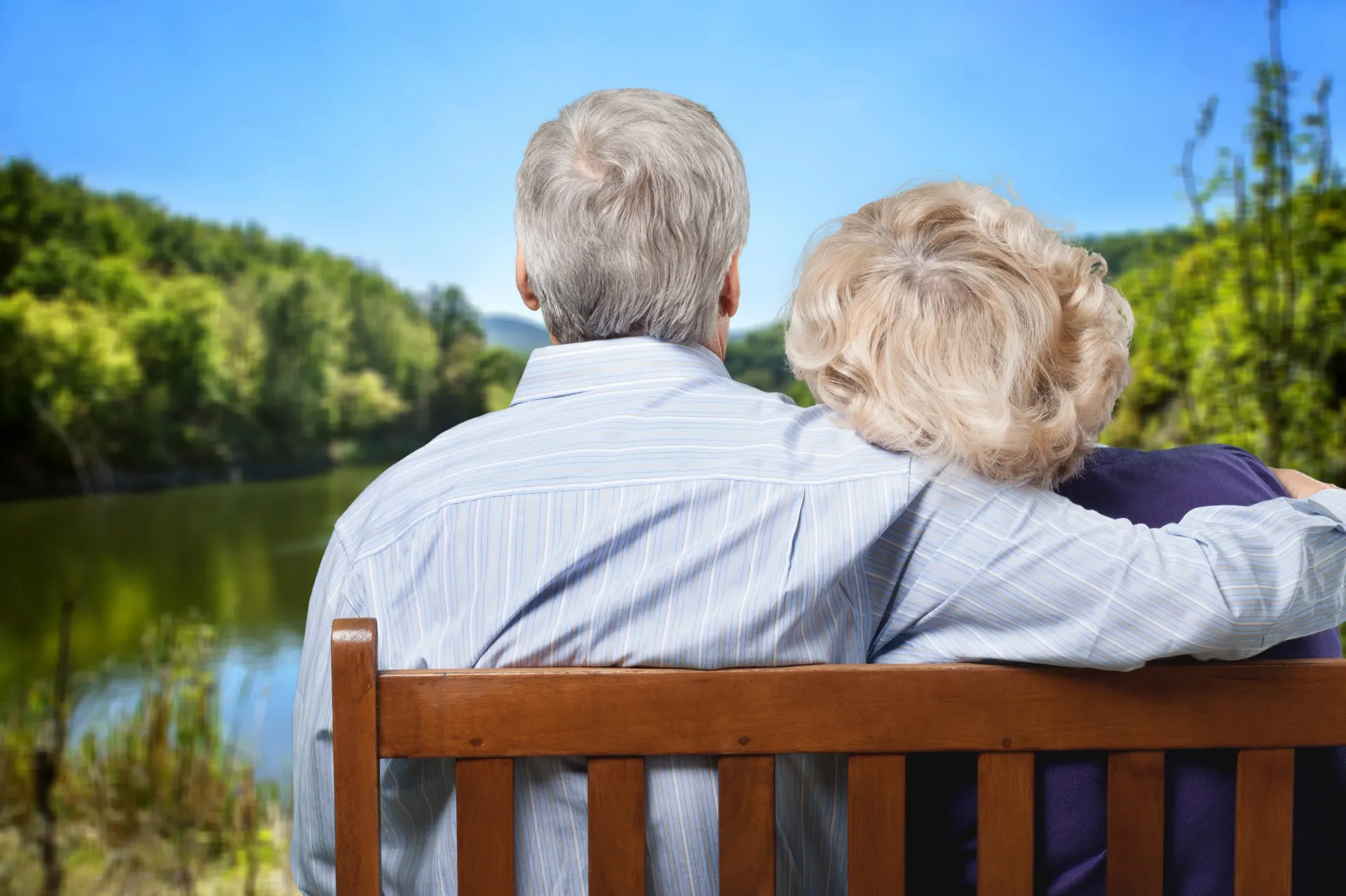 an elderly couple is sitting on a bench looking at a lake .