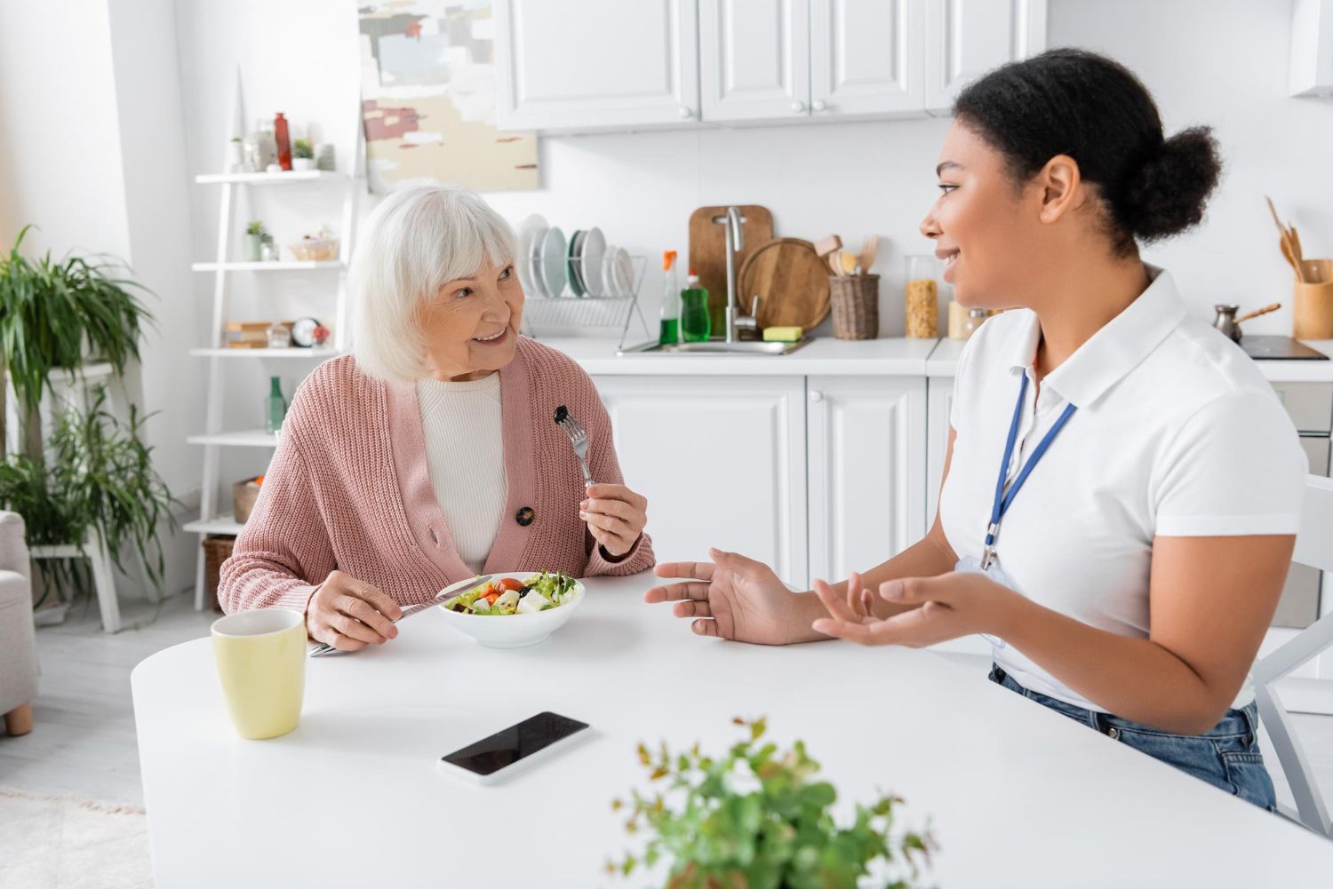 Two women talking at a kitchen table with food and a smartphone nearby
