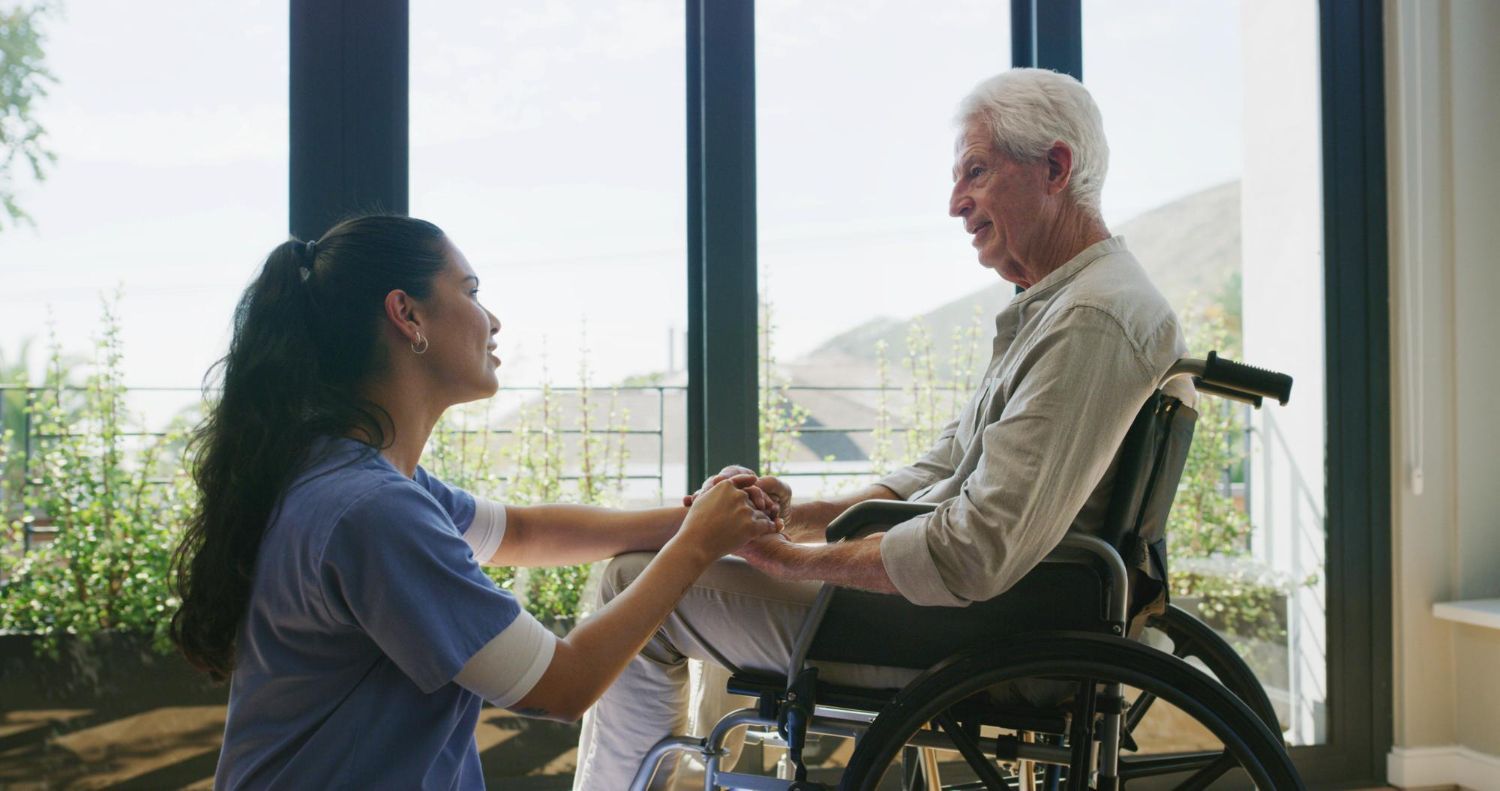 Woman holding hands with a smiling man in a wheelchair by a large window