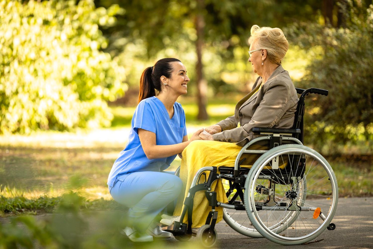 Caregiver kneels beside a person in a wheelchair outdoors, speaking and smiling in a sunny park.