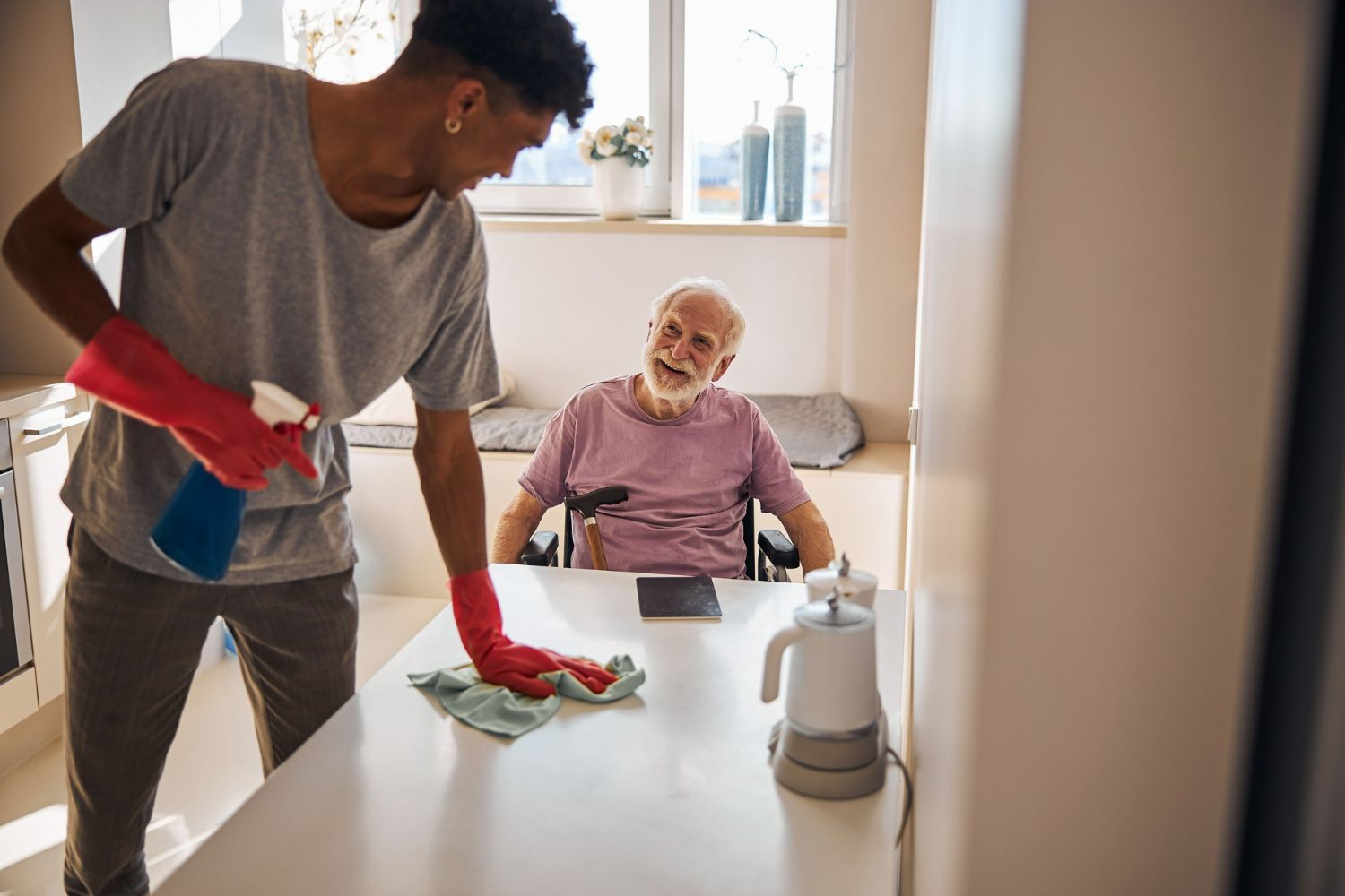 Man wiping a table while another man sits in a wheelchair and watches from a sunny kitchen.