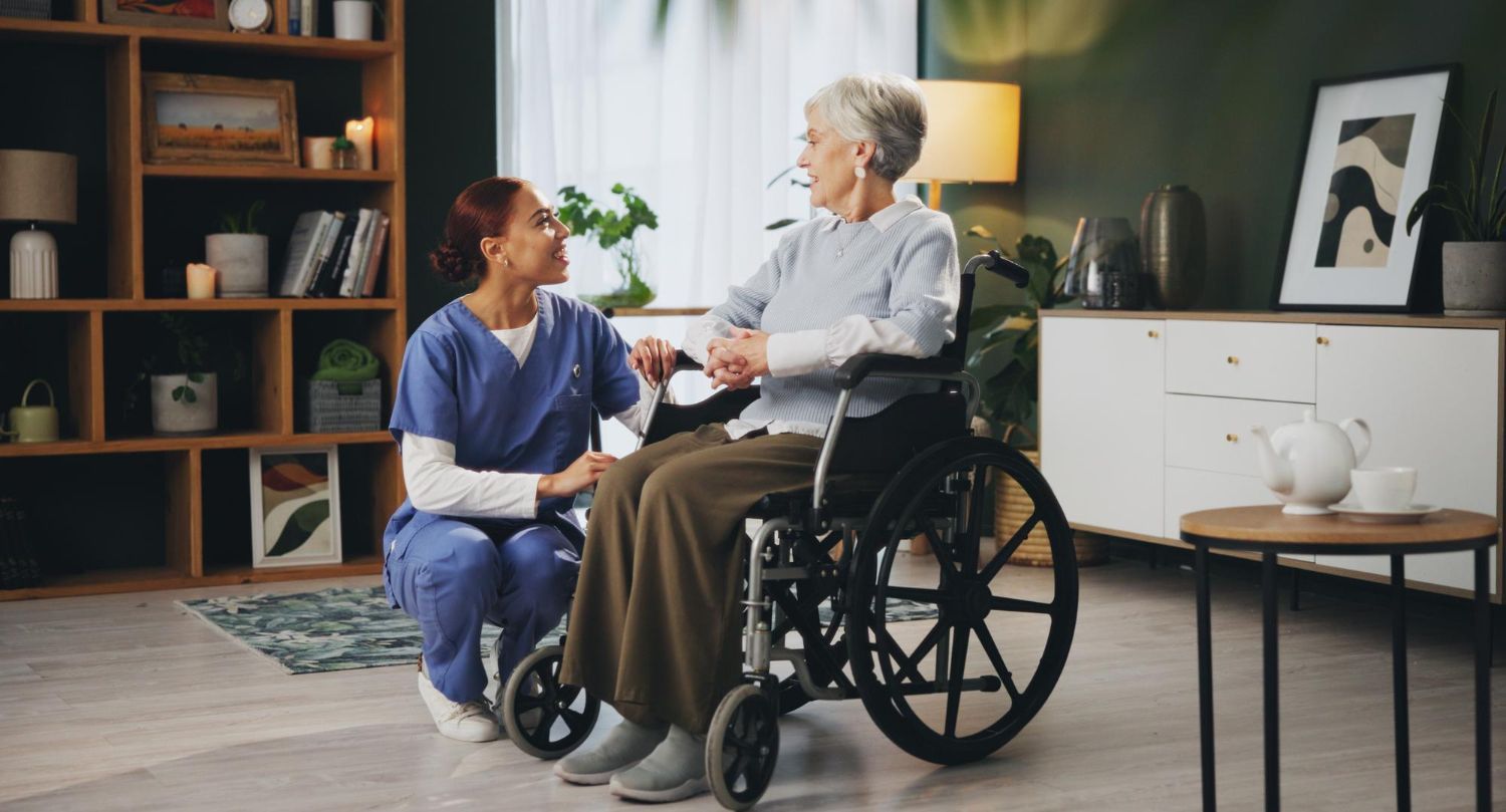 Caregiver kneels beside an older adult in a wheelchair in a cozy living room.