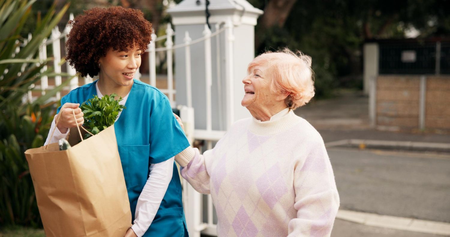 Two people walking outdoors, smiling and chatting, with a grocery bag and leafy greens visible.