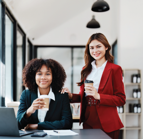Two coworkers in a bright office, one seated with a laptop and coffee, one standing in a red blazer holding a cup.