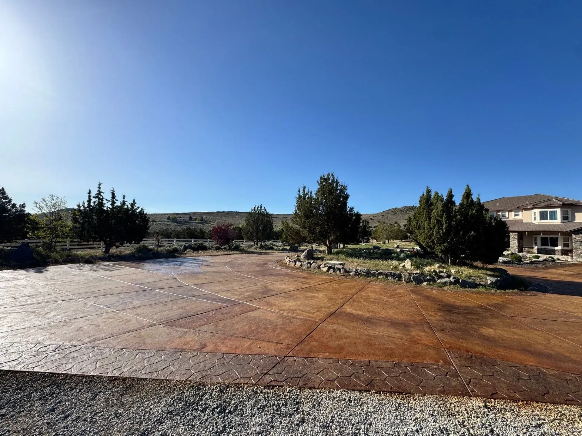 Wide gravel driveway beside a house, with trees and shrubs under a clear blue sky.
