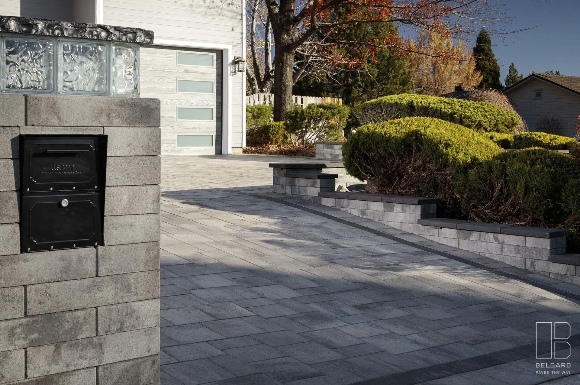 Modern house driveway with paver stones, stone mailbox, and landscaped shrubs in sunlight
