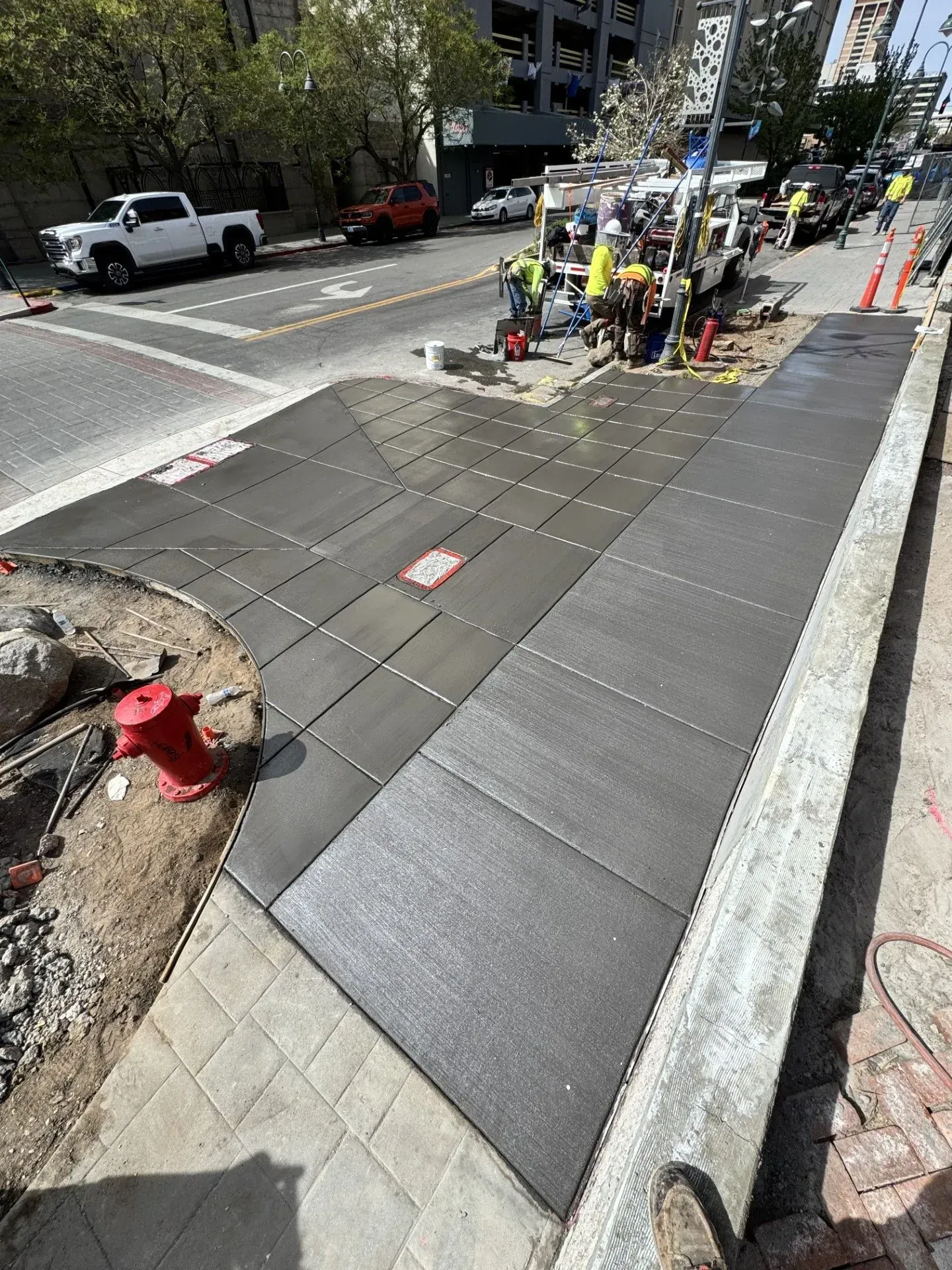 Sidewalk under repair with workers, orange cones, and exposed pavement along a city street.