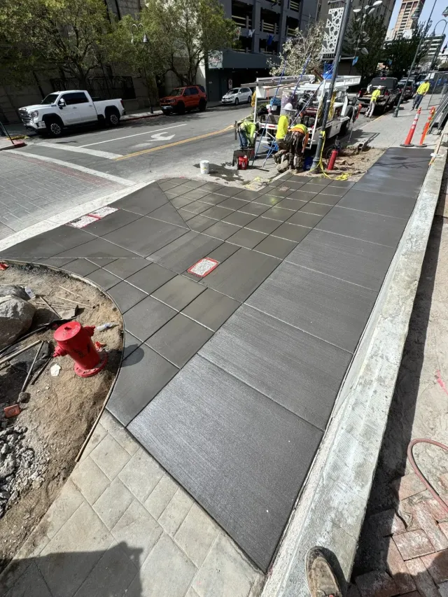 Sidewalk construction crew repairing a curb ramp with fresh gray pavers beside a city street.