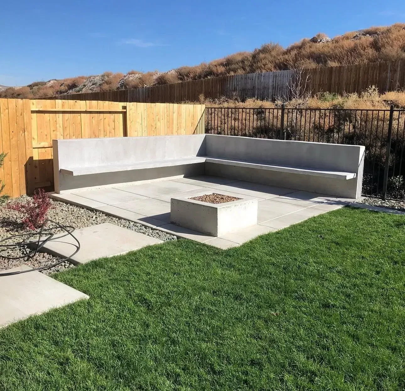 Outdoor patio with built-in gray concrete bench seating, fire pit, and artificial grass beside a wooden fence.